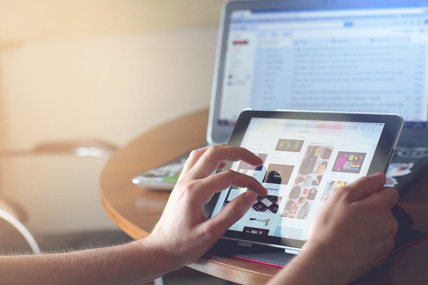 Person nutzt ein Tablet mit einer App, im Hintergrund ein Laptop mit geöffnetem E-Mail-Postfach