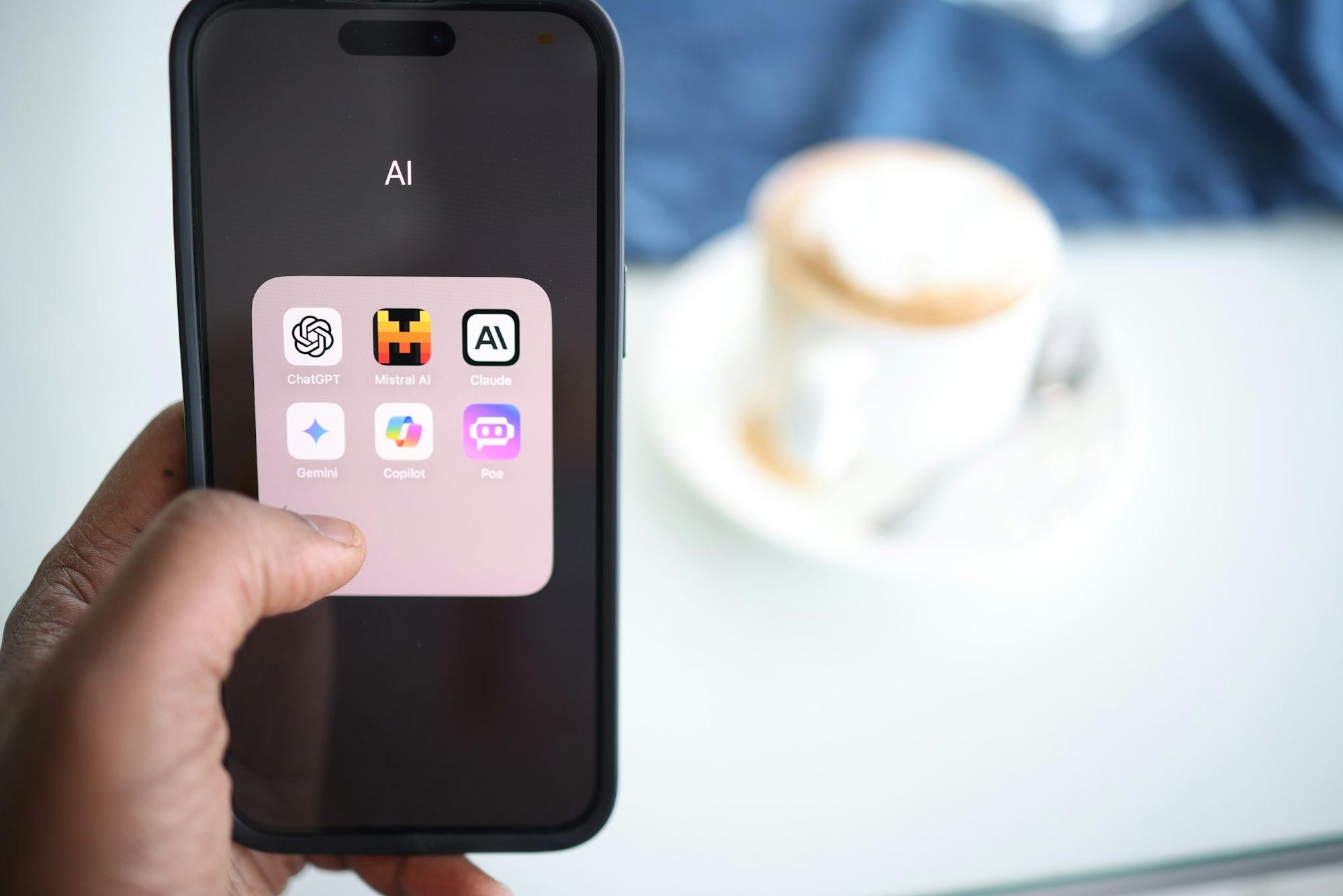A hand holding an iPhone whose home screen shows an "AI" folder with the app icons of ChatGPT, Mistral AI, Claude, Gemini, Copilot and Poe
