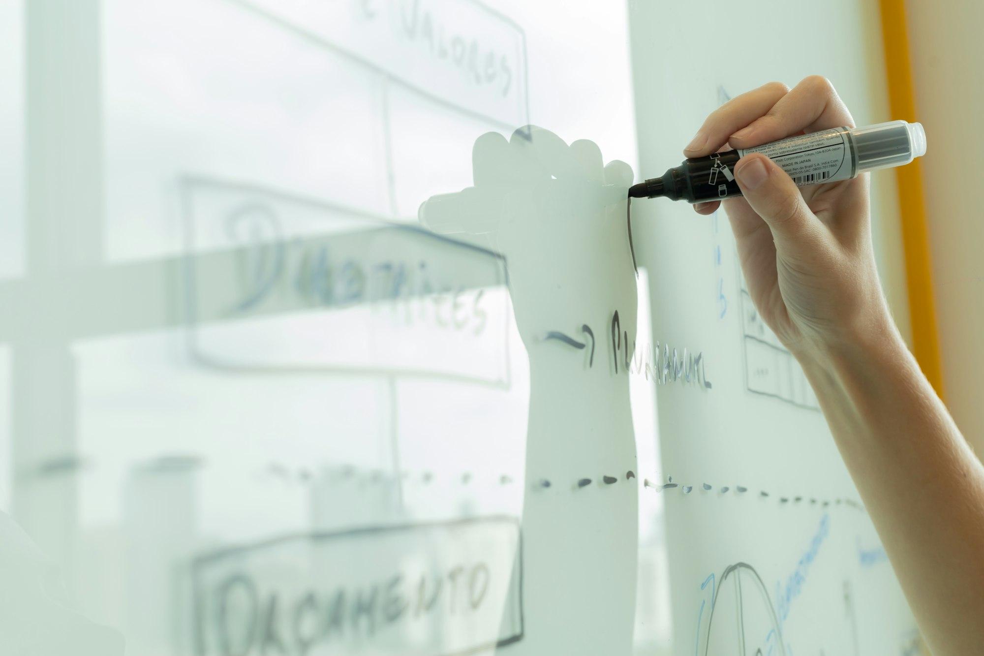 A hand drawing a diagram of boxes and connecting lines on a whiteboard with a marker