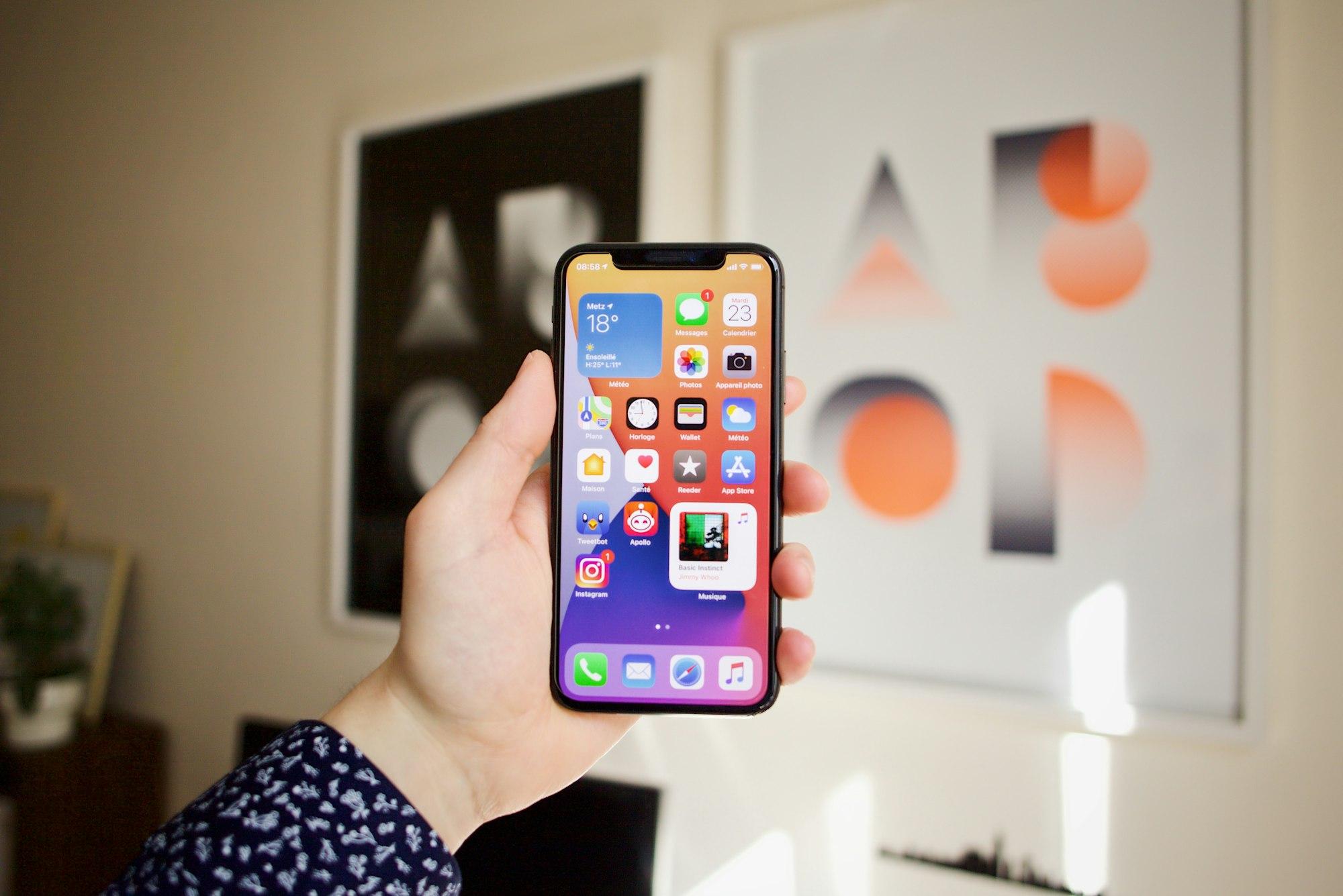 A hand holding an iPhone whose home screen shows app icons plus a weather and a music widget
