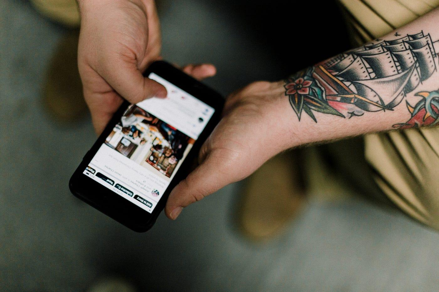 Eine Hand hält ein Smartphone mit geöffneter Social-Media-App, am anderen Arm ist ein Schiffs-Tattoo zu sehen