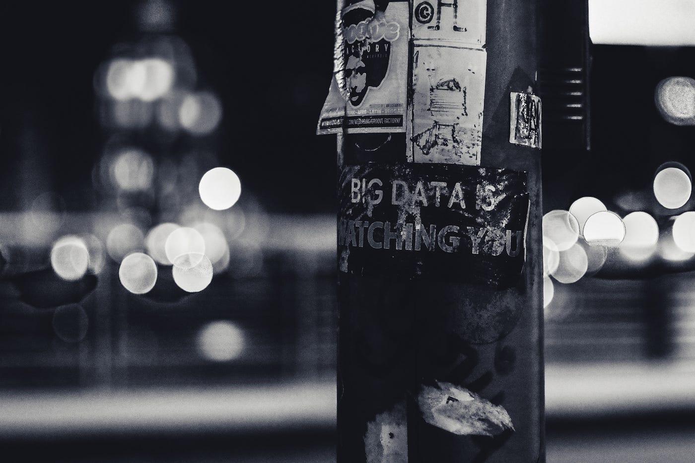 Ein Aufkleber an einer Laterne mit der Aufschrift 'Big Data is watching you' in einer nächtlichen Straßenszene in Schwarz-Weiß